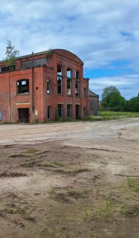 Former brick industrial building with tall windows beside open ground under a cloudy sky